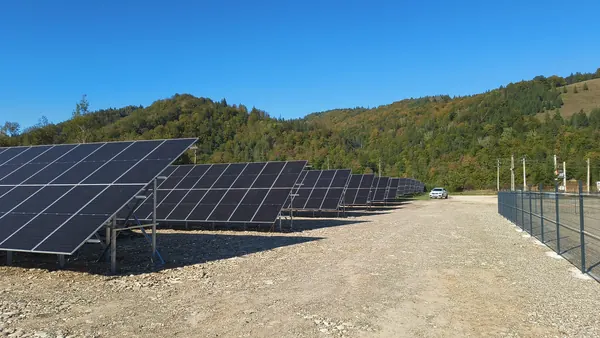 Parc fotovoltaic la sol, cu rânduri de panouri și dealuri împădurite